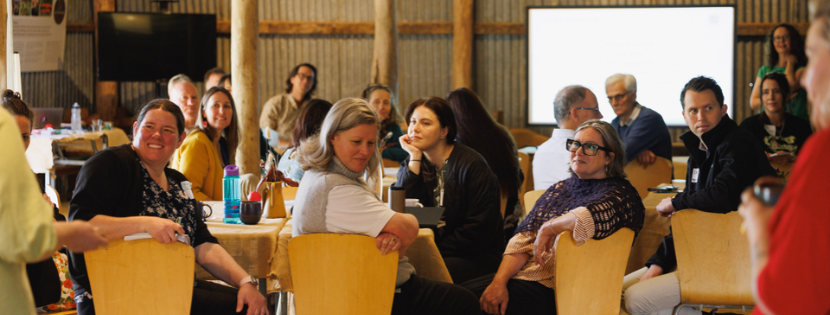 Group of people seated at round tables in a rustic indoor setting, attentively listening to a speaker off camera.
